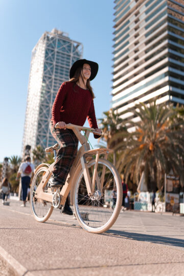 Ground level of merry young woman in casual outfit and hat smiling and riding eco friendly bicycle on pavement against skyscrapers and blue sky in tropical city in daytime.
